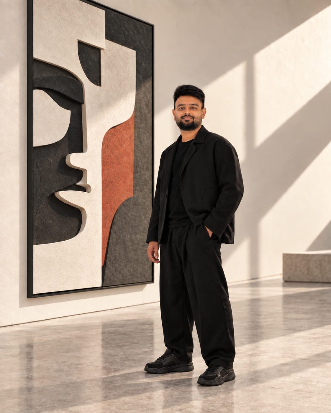 Deep Sata standing beside a large relief of a face in black, cream and rust, in a daylit gallery
